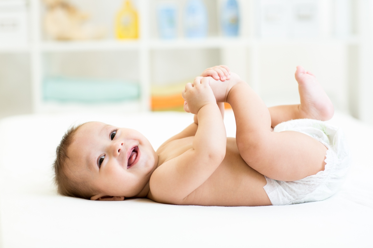 Lachendes Baby liegt auf einem weißen Bettlacken und hält siene Füße fest